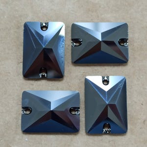 May include: Four rectangular, faceted, gray crystal rhinestones with silver-tone metal loops. The crystals have a geometric design with multiple facets, reflecting light. These decorative crystals are suitable for various craft projects.