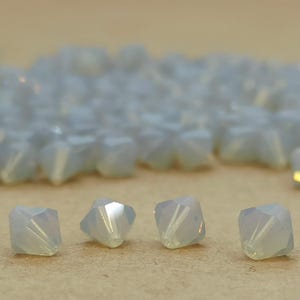 May include: A close-up of four iridescent white glass beads with a faceted, pyramid shape. The beads are arranged in a row on a brown surface.