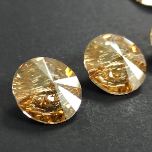 May include: Two round, faceted, amber-colored crystal rhinestones. The crystals have multiple facets that reflect light, creating a sparkling effect. The rhinestones are set against a dark background.