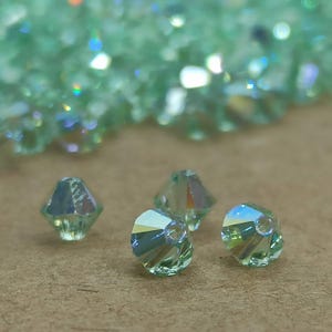 Puede incluir: Primer plano de cuatro cuentas de cristal verde claro con un diseño facetado. Las cuentas tienen un acabado brillante e iridiscente.