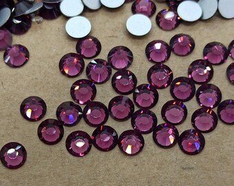 May include: Close-up of a collection of round, faceted, dark purple rhinestones. The rhinestones are scattered across a light brown surface, with some silver-backed, flat-backed stones visible.