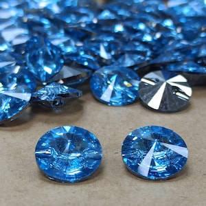 May include: A selection of light blue crystal buttons with a faceted design. The buttons are circular and reflect light due to their multiple facets. Some buttons are in focus, while others are blurred. The buttons are scattered on a neutral surface.