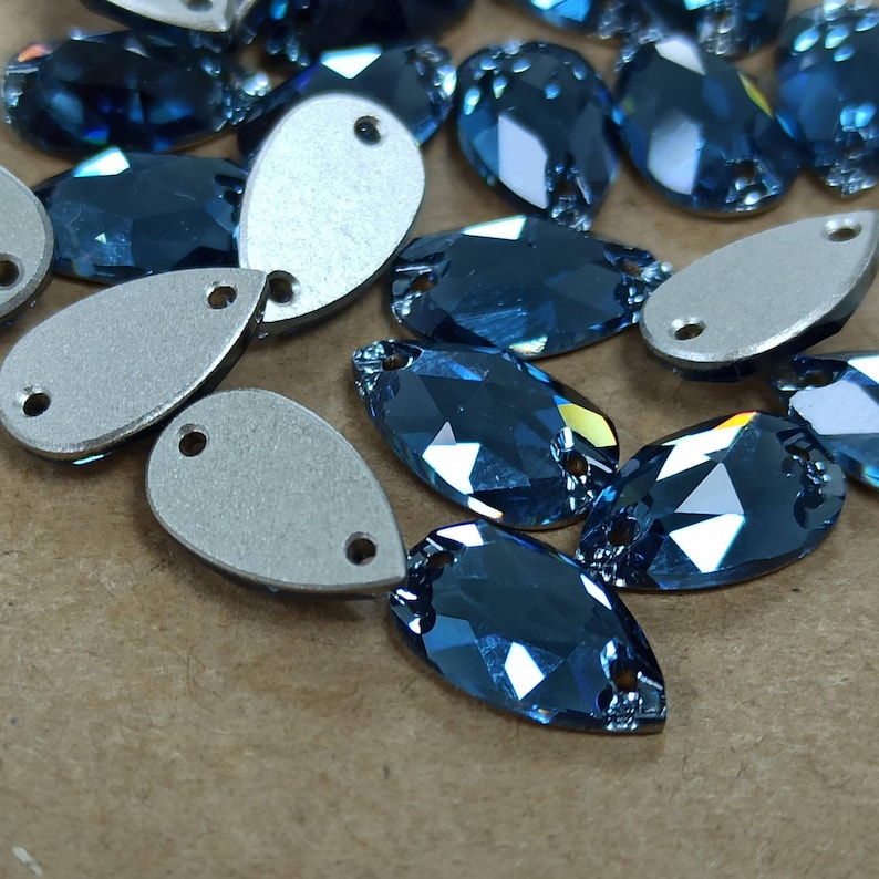 May include: Close-up of several blue teardrop-shaped rhinestones with silver backing. The rhinestones have multiple facets that reflect light. The silver backing has two small holes for sewing or attaching. These are ideal for jewelry making or embellishments.
