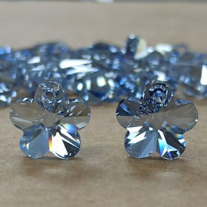 May include: Two light blue, faceted crystal flower-shaped beads. The beads have a five-petal design with a hole at the top for stringing. A blurred background shows more of the same beads.
