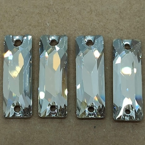 May include: Four clear, rectangular, faceted glass crystals arranged in a row. Each crystal features a polished surface with multiple geometric facets that reflect light and includes two small circular holes, one at the top and one at the bottom.