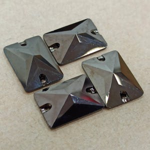 May include: Four black square-shaped rhinestones with a pyramid-shaped design. The rhinestones have a shiny, reflective surface.