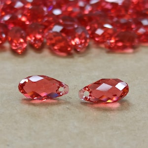 May include: Close-up of several faceted, teardrop-shaped red crystal beads. Two beads are in focus, showing a hole for stringing. The background is blurred with more red beads, suggesting a craft supply or jewelry component.