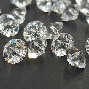 May include: A collection of clear, faceted crystal gemstones. The crystals have irregular shapes and multiple facets, reflecting light to create a sparkling effect. The gemstones are scattered on a dark surface, highlighting their brilliance.