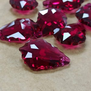 May include: Close-up of several faceted, deep red crystal beads. Each bead has multiple reflective white facets, creating a sparkling effect. The beads are irregularly shaped and appear to be made of glass or a similar material. The background is a neutral tan color.