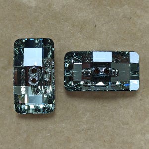 May include: Two rectangular, faceted buttons with a shimmering, iridescent finish. Each button has two holes for sewing. The buttons are a mix of clear and smoky gray, reflecting light. The buttons are on a neutral, tan background.