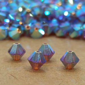 Peut inclure: Quatre perles de verre bleu irisé de forme en goutte facettées. Les perles ont un éclat métallique et sont disposées en rangée sur une surface brune.
