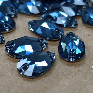 May include: Close-up of several faceted, teardrop-shaped blue rhinestones. Each gem has a flat back and a small hole at the top for stringing. The gems are arranged on a neutral-toned surface, reflecting light.