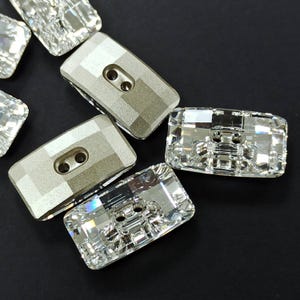 Puede incluir: Una colección de botones rectangulares con diseños de cristal facetado y metal plateado. Los botones tienen dos agujeros para coser y están dispuestos sobre una superficie negra. Los cristales reflejan la luz, creando un efecto brillante.