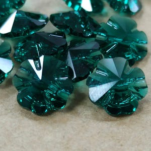 May include: Emerald green, faceted flower-shaped beads. The beads have a multi-faceted design, catching the light and creating a sparkling effect. The beads are clustered together, showcasing their intricate details and vibrant color. Ideal for jewelry making and crafts.
