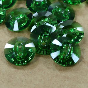 May include: A collection of faceted, emerald green buttons. Each button has a rounded shape with a central hole and white accents. The buttons are made of a clear, reflective material.