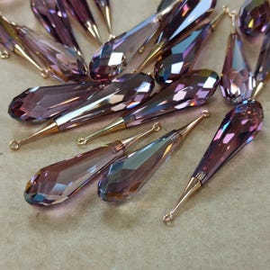 May include: A collection of faceted, teardrop-shaped crystal pendants in shades of purple and iridescent hues. Each pendant has a gold-colored metal cap and a small loop at the top for attaching to jewelry.