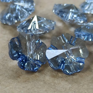 May include: A collection of light blue, flower-shaped crystal beads with faceted surfaces. Each bead features a central hole and a multi-faceted design that reflects light, creating a sparkling appearance across the translucent, pale blue glass material.