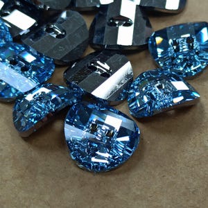May include: A collection of faceted, light blue glass buttons. The buttons have a unique shape with multiple facets that catch the light, creating a sparkling effect. Each button has two holes for sewing. The buttons are scattered on a brown surface.