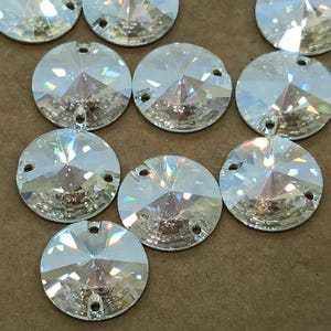 May include: A collection of clear, faceted, round rhinestones with three small holes for sewing or attaching. The rhinestones have a brilliant, sparkling appearance, reflecting light in multiple directions. These embellishments are ideal for adding sparkle to crafts and clothing.