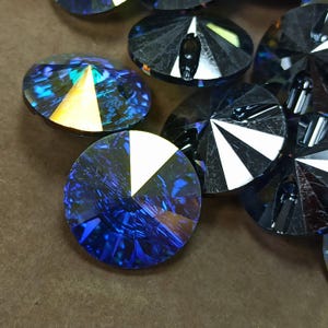 May include: A collection of circular, faceted glass buttons in a deep, iridescent blue hue. Each button features a distinct, reflective triangular segment that catches the light, creating a sharp contrast against the dark, multi-faceted surface.