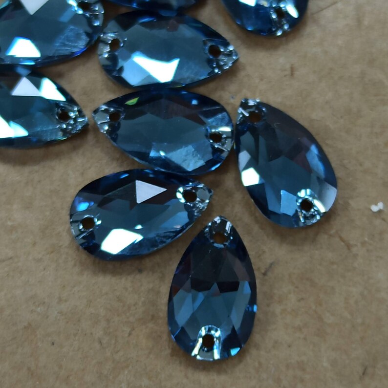 May include: Close-up of several blue teardrop-shaped rhinestones. Each rhinestone has a faceted surface and two small holes at the top for attaching. The rhinestones are scattered on a light brown surface.