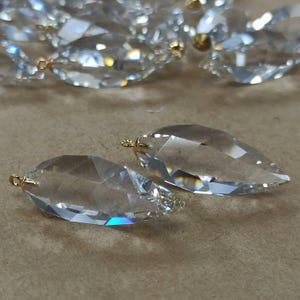May include: Two clear, faceted crystal teardrop pendants with gold-coloured metal findings. The crystals are highly reflective, catching and refracting light. The background is a neutral tan colour. Decorative crystals.