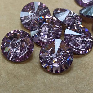 Peut inclure: Gros plan de plusieurs perles de cristal rondes, facettées, de couleur violet clair. Les perles ont une coupe complexe qui reflète la lumière, créant un effet scintillant. Les perles sont dispersées sur une surface neutre.