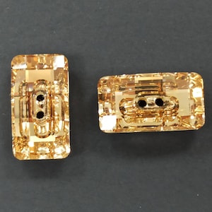 Könnte beinhalten: Zwei rechteckige, facettierte Knöpfe in Goldgelb. Jeder Knopf hat zwei Knopflöcher und ein dekoratives, strukturiertes Design. Die Knöpfe sind wahrscheinlich aus Glas oder Kristall.