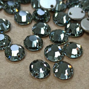 May include: A selection of grey, faceted rhinestones, round in shape, with a flat back and two small holes. The rhinestones have a reflective, sparkling appearance. Some are silver-coloured.