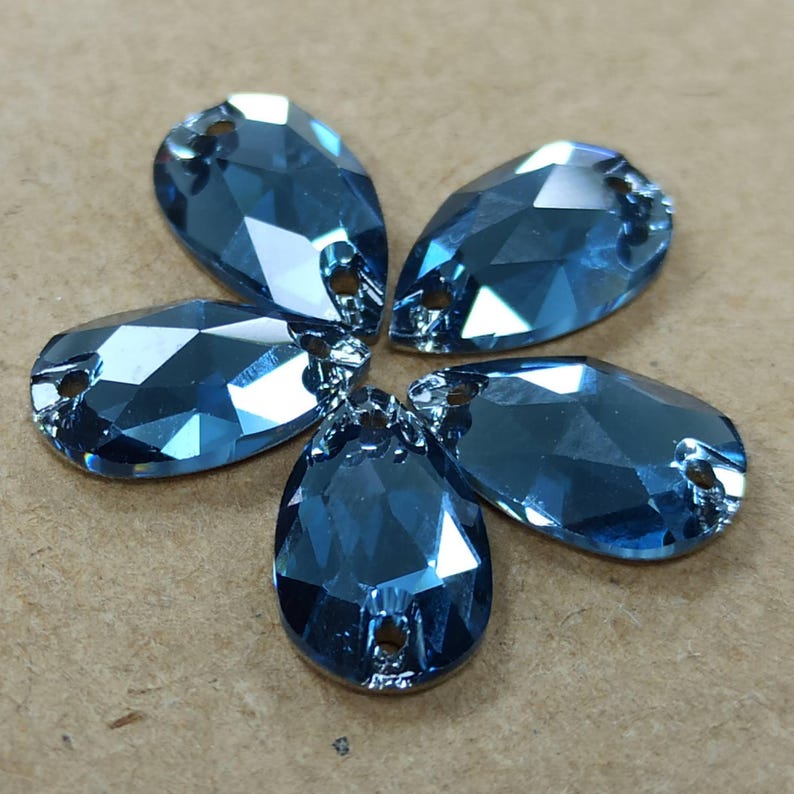 May include: Five blue teardrop-shaped rhinestones arranged in a flower-like pattern. Each rhinestone has a faceted surface and two small holes at the top for attaching to fabric or other materials. The rhinestones are a vibrant, clear blue color.