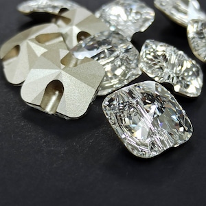 Puede incluir: Primer plano de varios pedrería de cristal transparente y plateado. Los cristales están facetados, reflejan la luz y tienen una variedad de formas, incluyendo diseños cuadrados y geométricos. El fondo es una superficie negra sólida.