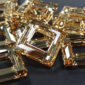 Puede incluir: Primer plano de varias gemas cuadradas de color marrón dorado cálido. Cada gema tiene un centro hueco y abierto y refleja la luz. Las gemas están dispersas sobre una superficie oscura, creando un efecto brillante.