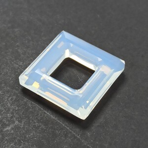Puede incluir: Una gema opalescente de forma cuadrada con un recorte cuadrado en el centro. La gema tiene un color azul pálido y blanco, con una superficie facetada que captura la luz. Probablemente se utiliza para la fabricación de joyas.