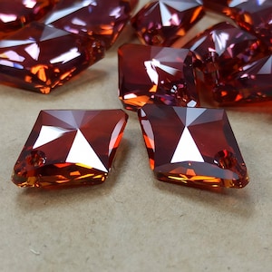 May include: Close-up of several faceted, reddish-orange crystal beads. The beads are diamond-shaped with a hole drilled through one corner. The crystals reflect light, creating bright highlights and shadows. The beads are likely for jewelry making.