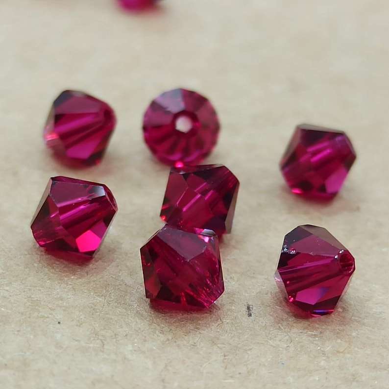 May include: A group of eight ruby red crystal beads, each with a faceted, square shape. The beads are scattered on a brown surface.