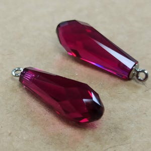 May include: Two faceted, teardrop-shaped, ruby-red crystal pendants with silver loops. The crystals are pointed and designed to catch the light. These pendants are likely for jewellery making or as decorative elements.