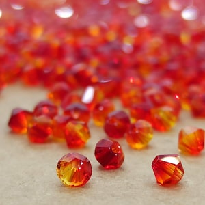 Puede incluir: Primer plano de cuentas de vidrio facetadas rojas y naranjas. Las cuentas están dispersas sobre una superficie neutra, con una mezcla de tonos rojo intenso y naranja vibrante. La luz atrapa las facetas, creando un efecto brillante. Ideal para la creación de joyas.