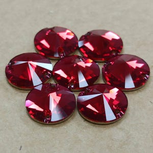 May include: Seven round, faceted, red crystal rhinestones with a flat back. The crystals have a brilliant shine and are arranged in a circular pattern. Each crystal has a small hole near the top for attachment.