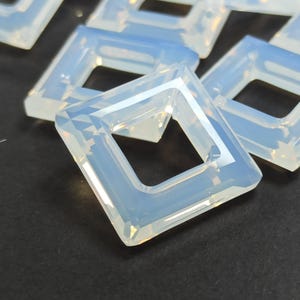 Può includere: Primo piano di diverse perline iridescenti a forma quadrata. Ogni perlina ha un foro quadrato al centro ed è sfaccettata per catturare la luce. Le perline sono di un colore traslucido e opalescente, con sfumature di blu e giallo.