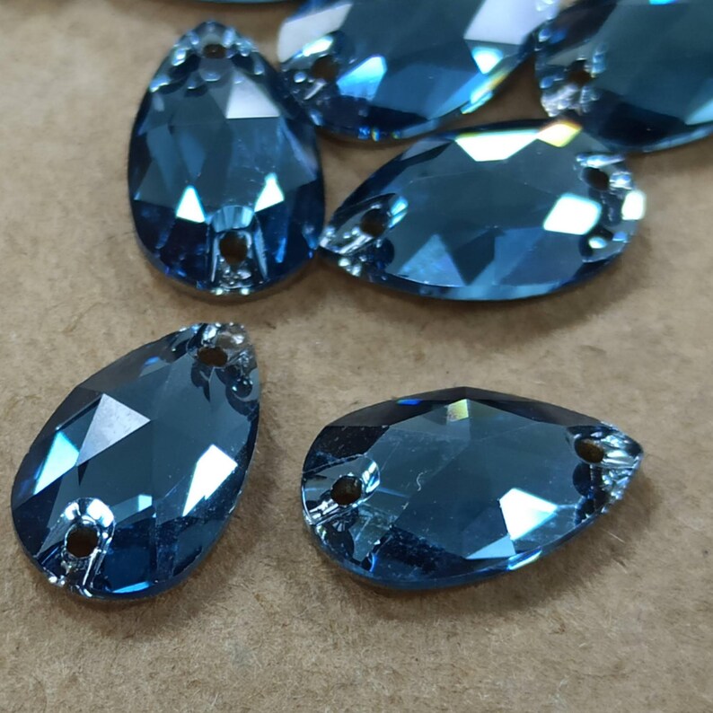 May include: Several faceted, teardrop-shaped blue gemstones. Each gem has a flat back and two small holes near the top. The gems are a deep, translucent blue, reflecting light with multiple facets. These could be used for jewelry making or craft projects.