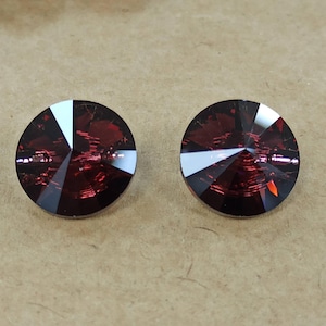 May include: Two round, faceted, dark red crystal stones. Each stone has a white triangular facet. The crystals are set against a neutral, light brown background. These crystals could be used for jewellery making or other craft projects.
