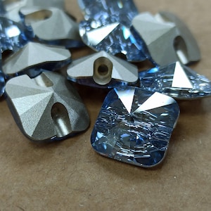 May include: Close-up of several light blue square crystal buttons with faceted surfaces. Some buttons have a silver metal backing with a hole. The buttons are scattered on a brown surface.