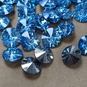 Può includere: Una collezione di perline di cristallo sfaccettate, rotonde, blu chiaro. Alcune perline sono di un colore argento brillante e riflettente. Le perline sono sparse su una superficie dai toni neutri. Adatto per la creazione di gioielli e progetti artigianali.