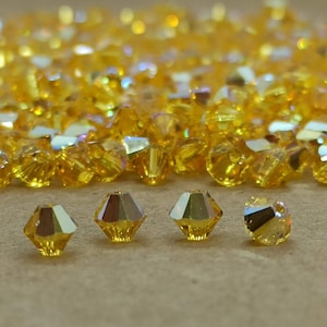 May include: A close-up of a pile of small, golden yellow, faceted glass beads. The beads are arranged in a random pattern and have a shiny, iridescent finish.