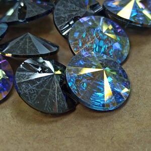 Op de afbeelding: Close-up van verschillende iriserende, gefacetteerde kristallen strass-steentjes. De kristallen zijn rond met meerdere facetten en reflecteren een spectrum aan kleuren, waaronder blauw, geel en paars. De kristallen liggen verspreid op een bruin oppervlak.