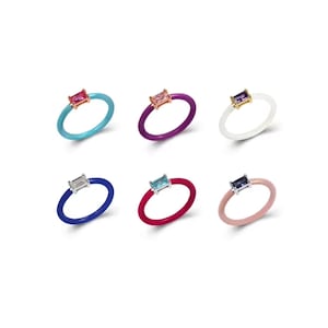 Puede incluir: Seis anillos de silicona coloridos con una pequeña piedra rectangular engastada en un engaste de oro. Los anillos son de color turquesa, morado, blanco, azul, rojo y rosa.