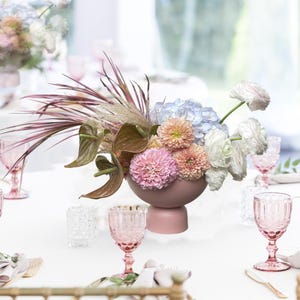 Puede incluir: Elegante mesa decorada con un centro floral de flores rosas, azules y blancas en un jarrón malva. Copas rosas y sillas doradas rodean el mantel blanco. Un arreglo sofisticado para un evento especial.