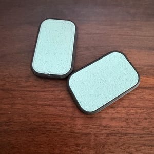May include: Two rectangular, mint-green sponges with dark gray borders are displayed on a wooden surface. The sponges have a textured appearance and a rounded rectangular shape.