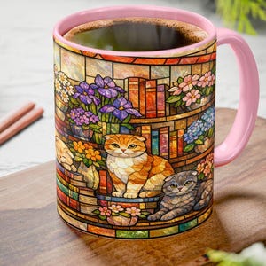 Puede incluir: Una taza de caf&eacute; de cer&aacute;mica con asa y borde rosas, con un dise&ntilde;o de vidriera. El dise&ntilde;o incluye dos gatos, flores y libros. La taza est&aacute; llena de caf&eacute; y se encuentra sobre una tabla de cortar de madera.