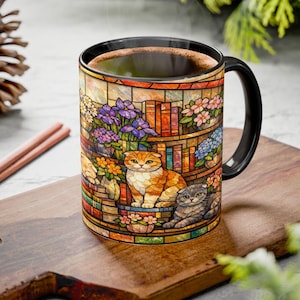 May include: A ceramic coffee mug with a black handle and rim, featuring a stained-glass design of a bookshelf, flowers, and two cats. The mug is filled with coffee and sits on a wooden cutting board.